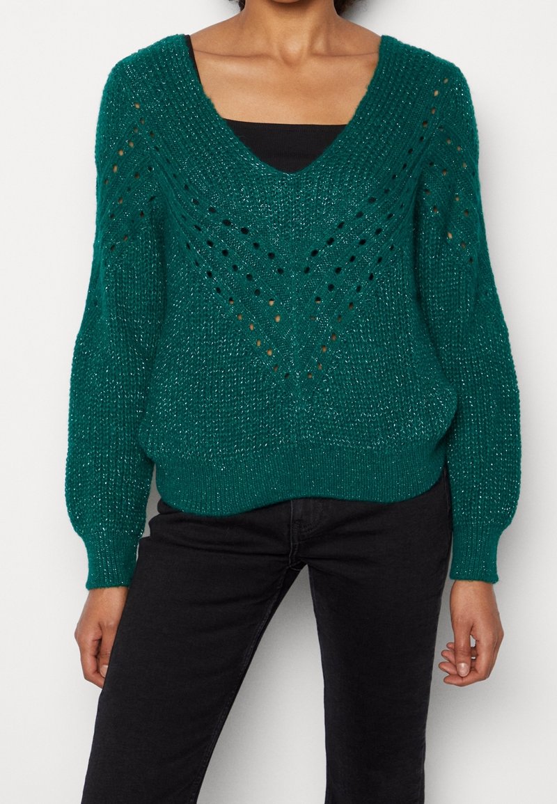 Pull tricoté sarcelle avec un col en V, présentant des motifs à points ouverts et des poignets côtelés. Porté sur un haut noir, associé à un pantalon noir.