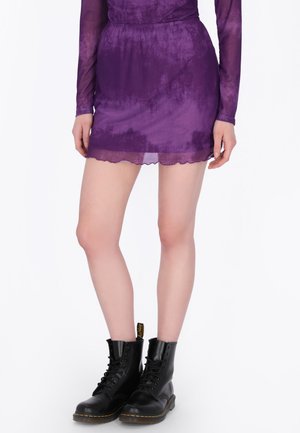 Frau trägt ein durchsichtiges lila Minikleid mit langen Ärmeln und schwarze Schnürstiefel auf weißem Hintergrund.