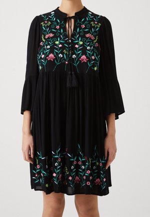 Robe noire longueur genou avec broderie florale colorée sur la poitrine et l'ourlet, manches trois-quarts et lien avec pompons au col.