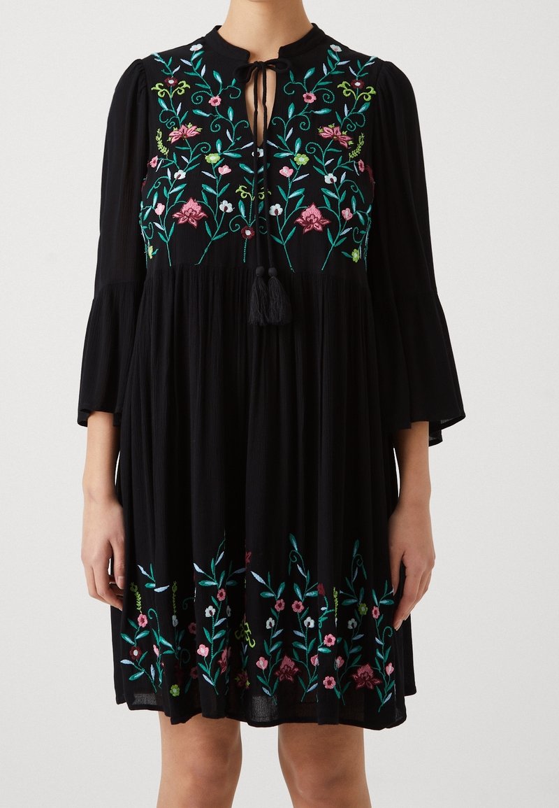 Robe noire longueur genou avec broderie florale colorée sur la poitrine et l'ourlet, manches trois-quarts et lien avec pompons au col.