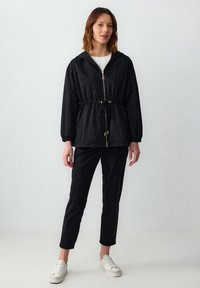 Zwarte lichtgewicht jas met een trekkoord in de taille, een ritssluiting aan de voorkant en een capuchon. Gepaard met zwarte broeken en witte sneakers, tegen een neutrale achtergrond.