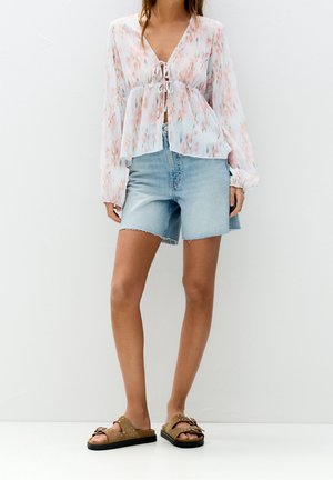 Woman wearing a sheer floral long-sleeve blouse tied at the front, light denim shorts, and brown studded sandals standing against a white wall.