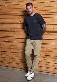 Námořnické tričko s logem, béžové kalhoty, bílé a hnědé tenisky; muž se opírá o dřevěnou zeď, detailní tetování viditelné na pažích.