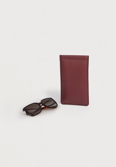 Lunettes de soleil noires à monture carrée avec branches marron à côté d'un étui à lunettes en cuir rectangulaire vertical bordeaux sur fond blanc.