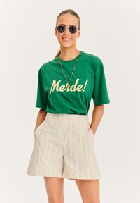 Zelené tričko s nápisom "Merde!" v žltom písmene, skombinované s béžovými pruhovanými šortkami. Slnečné okuliare a náhrdelník z korálkov dotvárajú vzhľad.