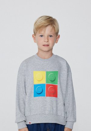 Blondýnský chlapec, který nosí šedou mikinu se čtyřmi barevnými ikonami LEGO kostek v žluté, zelené, modré a červené na přední straně.