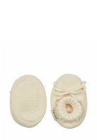 Szydełkowe buciki dla niemowląt w kremowym kolorze, o okrągłym kształcie, z elastycznym otworem i kokardką oraz strukturalnym wzorem. Metka z logo widoczna z boku.