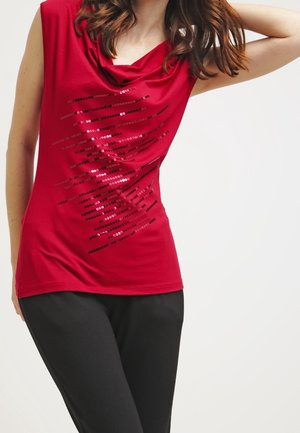 Kvinde iført rød ærmeløs top med sorte palietdiagonale striber, parret med sorte bukser, arm løftet bag hovedet.