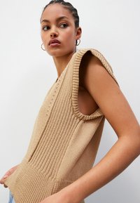 Beige kudumaterjalist ilma varrukateta topp, millel on soonikstruktuur, laiad käemised ja V-kaelus. Nähtavad õmblused ja lõdvestunud lõige.