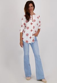 Witte blouse met rode bloemenborduursels, gecombineerd met lichtblauwe flared jeans en lichtroze espadrilles, staand voor een neutrale achtergrond.
