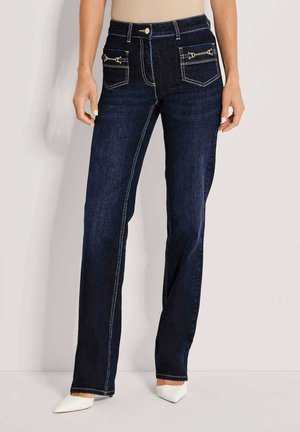 Vrouw draagt donkerblauwe rechte jeans met gouden kettingdetail op de voorzakken en witte pumps met puntige neus tegen een lichte achtergrond.