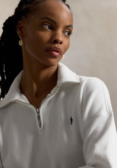 Weißer Pullover mit Viertel-Reißverschluss aus weichem Stoff, mit geripptem Kragen, silbernem Reißverschluss und einem kleinen dunkelblauen Logo auf der linken Brust.