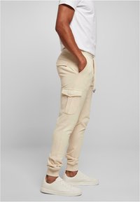 Beige cargo joggers with side pockets, tapered cuffs, and drawstring waist, paired with white sneakers and a plain white t-shirt.