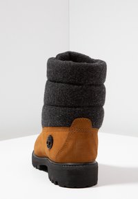 Bottine en daim marron avec un revers en feutre gris foncé, texturée de plis horizontaux, arborant un logo circulaire noir sur le côté et une semelle robuste.