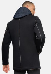 Black coat featuring a detachable navy quilted hood, contrasting stitching, and a structured, tailored fit. Smooth fabric with a textured finish.