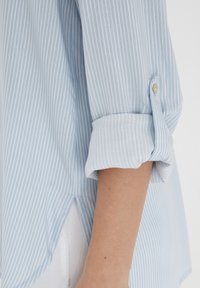 Light blue striped shirt with rolled-up sleeves, featuring a button detail and soft, textured fabric. Side slit at the bottom hem.
