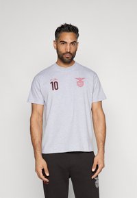 Γκρι βαμβακερό μπλουζάκι με κοντά μανίκια, που διαθέτει κόκκινο έμβλημα και αριθμό 10 στο μπροστινό μέρος. Το μοντέλο φοράει ταιριαστά σκούρα παντελόνια.