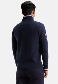 Pull-over en tricot marine avec un col haut, des poignets côtelés et une accent rayé sur la manche gauche, présentant une texture lisse et une coupe ajustée.