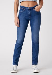 Žena v slim-fit středně vysokých modrých džínách s rukou v kapse a bílých teniskách, v croppované denimové blůze, stojící na jednobarevném pozadí.