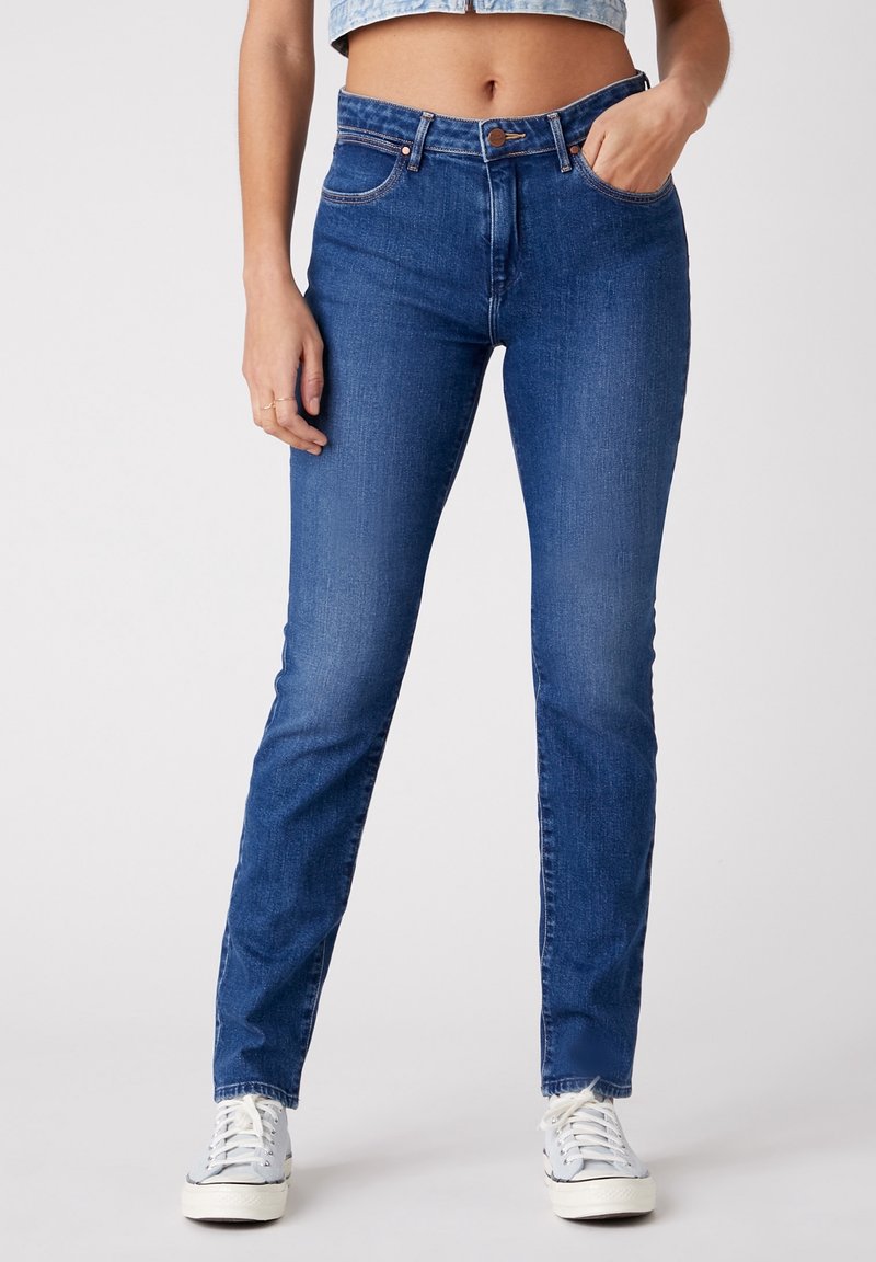 Žena v slim-fit středně vysokých modrých džínách s rukou v kapse a bílých teniskách, v croppované denimové blůze, stojící na jednobarevném pozadí.