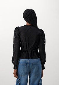 Schwarze Bluse mit strukturiertem Muster, voluminösen Ärmeln und elastischem Bund, kombiniert mit hellblauen Jeans-Cargohosen.