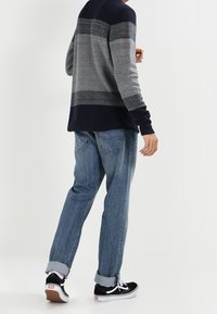 Person som bär en grå och svart randig tröja, uppvikta blå jeans och svarta Vans-sneakers, står med ryggen mot kameran.