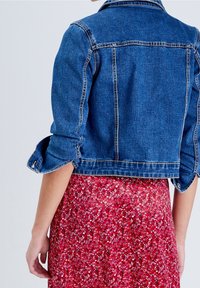 Blå denimjacka med en kortad design och uppkavlade ärmar, med gula stickningar och en klassisk krage, kombinerad med en röd blommig kjol.