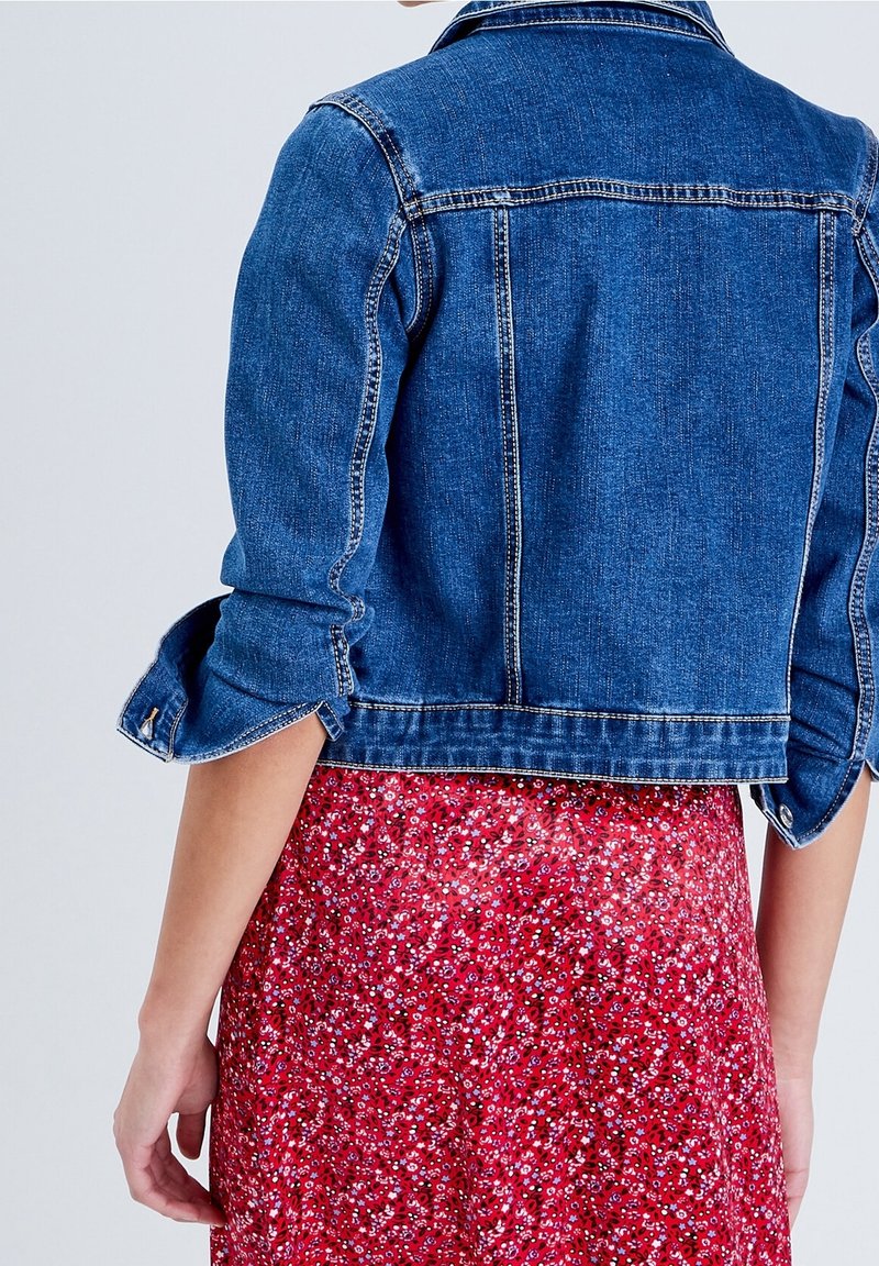 Blå denimjacka med en kortad design och uppkavlade ärmar, med gula stickningar och en klassisk krage, kombinerad med en röd blommig kjol.