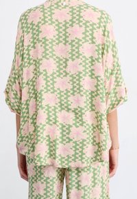 Blouse légère verte avec un motif floral rose, dotée d'une coupe ample, de larges manches et d'une fermeture à boutons au dos.