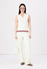 Top sem mangas branco canelado com decote riscado em vermelho e branco, combinado com calças brancas largas e calçado com padrão de leopardo, segurando uma bolsa branca.