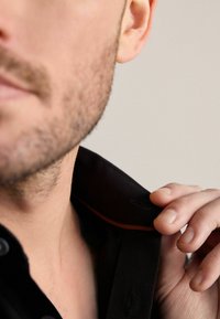 Nahaufnahme eines schwarzen Hemdkragens mit orangefarbenen Nähten, teilweise sichtbar. Eine Hand greift den Kragen und zeigt die Stofftextur sowie Designdetails.