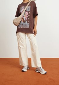 Camiseta gráfica marrón con diseño floral; pantalones de pierna ancha color crema; zapatillas deportivas grises con acentos en azul marino; bolso cruzado beige.