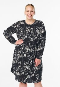 Svart blommönstrad klänning med långa ärmar, knyts i midjan och har en asymmetrisk fåll. Har vita blommönster och rund halsringning.