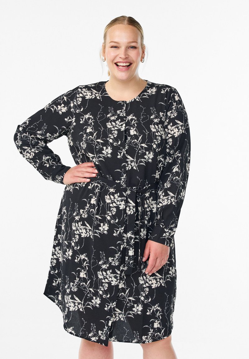 Robe noire à fleurs avec des manches longues, une taille nouée et un ourlet asymétrique. Présente des motifs floraux blancs et un col rond.