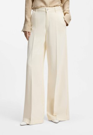 Kobieta ubrana w kremowe spodnie z wysokim stanem i szerokimi nogawkami, z przodem z zakładkami, bocznymi kieszeniami i zapięciem na guzik, w połączeniu z białymi butami na szpic.