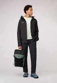 Veste softshell noire, sweat à capuche gris zippé, chemise vert clair, pantalon noir, chaussures bleues et un sac à dos bicolore avec un logo.