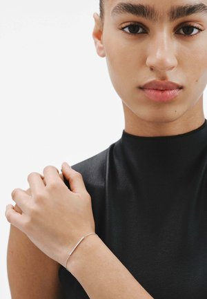 Pulsera de plata con una barra delgada y alargada sobre una delicada cadena, llevada en una mano sobre un fondo de tela negra texturizada.