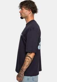 Sort oversized T-shirt med korte ærmer, lige kant og lys blå grafisk detalje på ryggen; modellen vist fra siden.