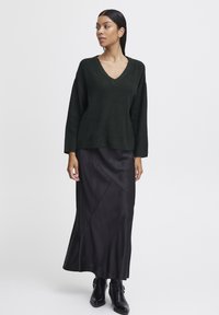 Pull en maille côtelée vert foncé avec un col en V, associé à une jupe maxi en satin noir avec une couture diagonale et des bottines noires.
