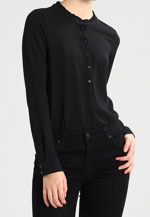 Kobieta ubrana w czarną, czarną, długą koszulę z guzikami i falbankowym kołnierzem oraz czarne spodnie z wysokim stanem z subtelnymi paskami.