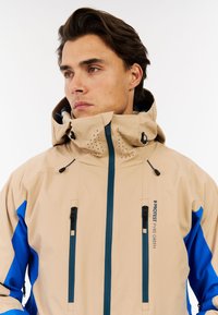 Bežinė ir mėlyna atspari vandeniui striukė su aukštu apykakliu, užtrauktuku užsegamomis kišenėmis ir ventiliacinėmis skylėmis. Turi logotipą ant krūtinės ir reguliuojamus užsegimus.