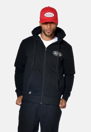 Homme portant un hoodie noir zippé avec des poches avant et un logo blanc, une chemise blanche en dessous, et une casquette rouge avec le même logo.