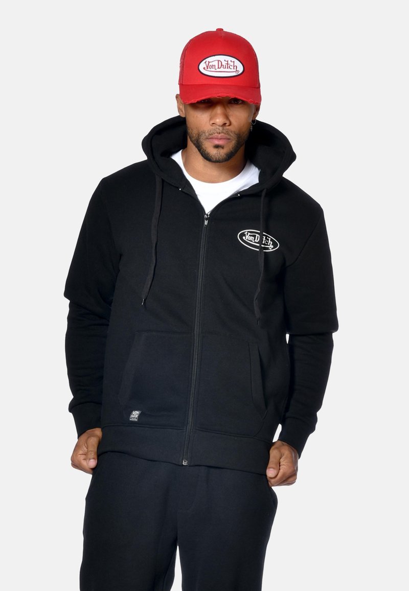 Uomo che indossa una felpa nera con zip, con tasche frontali e un logo bianco, una maglietta bianca sotto e un cappellino rosso con lo stesso logo.