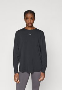 Μαύρο μπλουζάκι με μακρύ μανίκι και στρογγυλή λαιμόκοψη, από λεία ύφανση, με λευκό λογότυπο Nike στο στήθος, συνδυασμένο με γκρίζες κολάν.