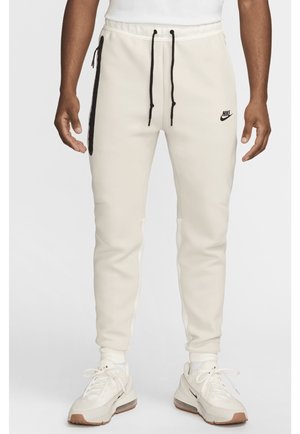 Man die off-white Nike joggers draagt met een zwart zijritszakje en aantrekkoorden, gecombineerd met witte Nike sneakers en sokken.