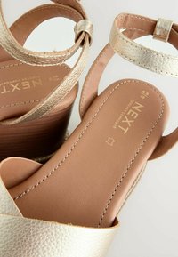 Arany bőrből készült platform szandál bokapánttal, texturált felülettel, barna párnázott talpbéléssel és fa hatású blokk sarokkal.