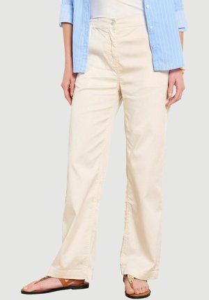 Sieviete, tērpusies bēša krāsas platās biksēs, zilā svītrainā kreklā ar pogām un brūnās plakanās sandālēs, stāv pret vienkāršu gaišu fonu.