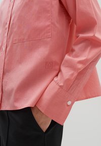 Chemise corail à manches longues avec fermeture à boutons, poche poitrine et logo brodé sur la manche. Tissu lisse et texturé avec une coupe ajustée.