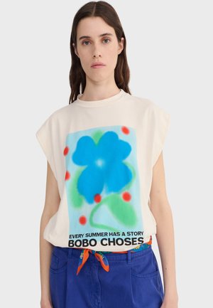 Femme portant un haut beige sans manches avec un grand motif de fleur bleue et des points rouges ainsi que le texte « Chaque été a une histoire Bobo Choses », pantalon bleu.