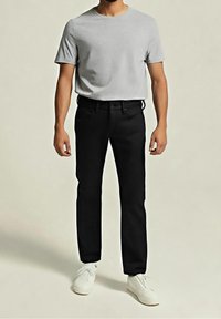 Homme portant un t-shirt gris clair à manches courtes, un pantalon noir et des baskets blanches, debout devant un fond uni clair.